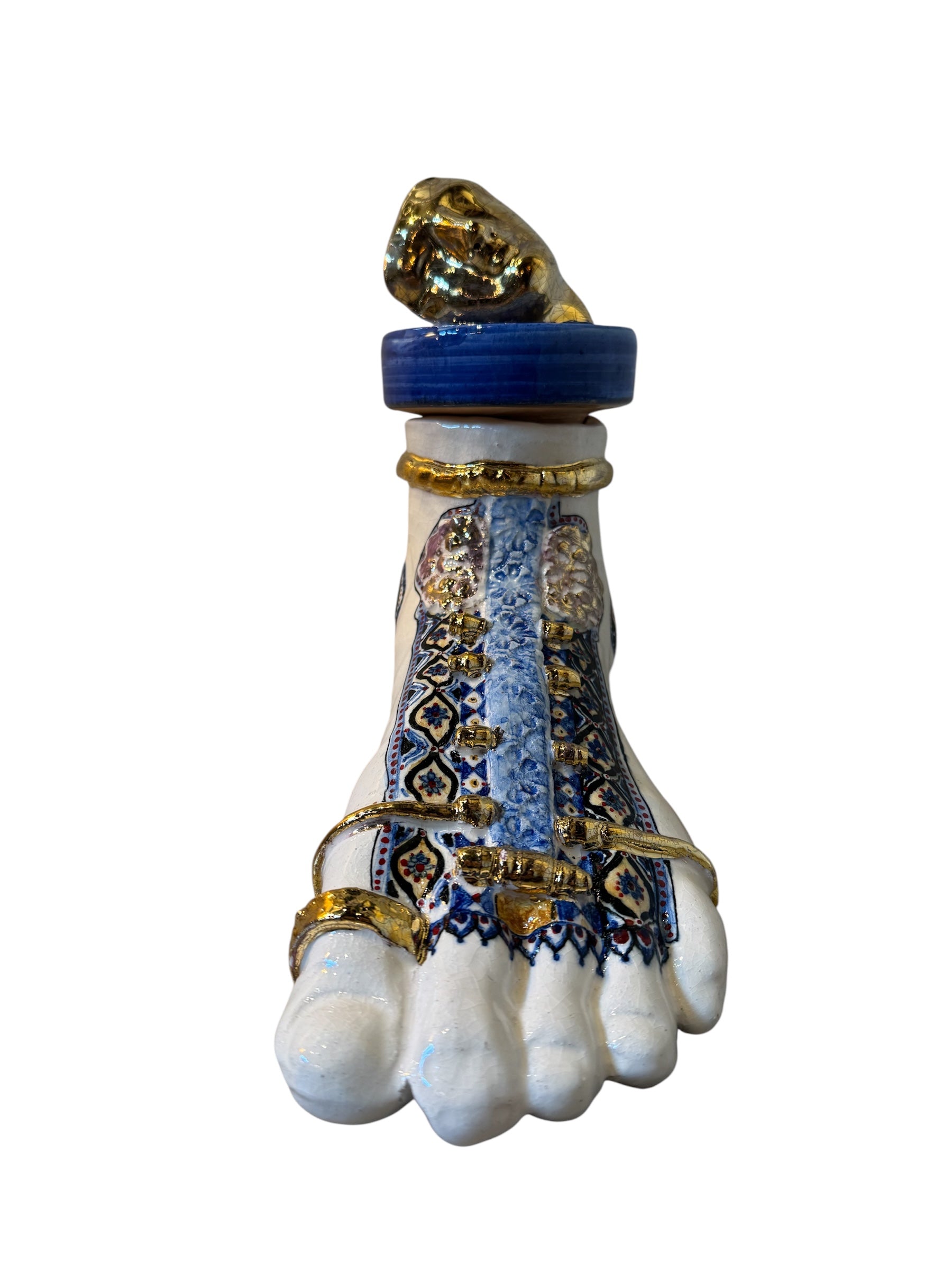 Close-up of a ceramic foot sculpture by Domenico Pinto. The foot is decorated with intricate hand-painted designs inspired by Apulian folklore and accented with 24KT gold. A stylized gilt mask sits atop the foot.