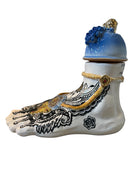 Handcrafted ceramic sculpture in the shape of a foot, decorated with intricate black and white patterns, 24KT gold accents, and topped with a blue lid featuring a gilt fish and head emerging from stylized waves.