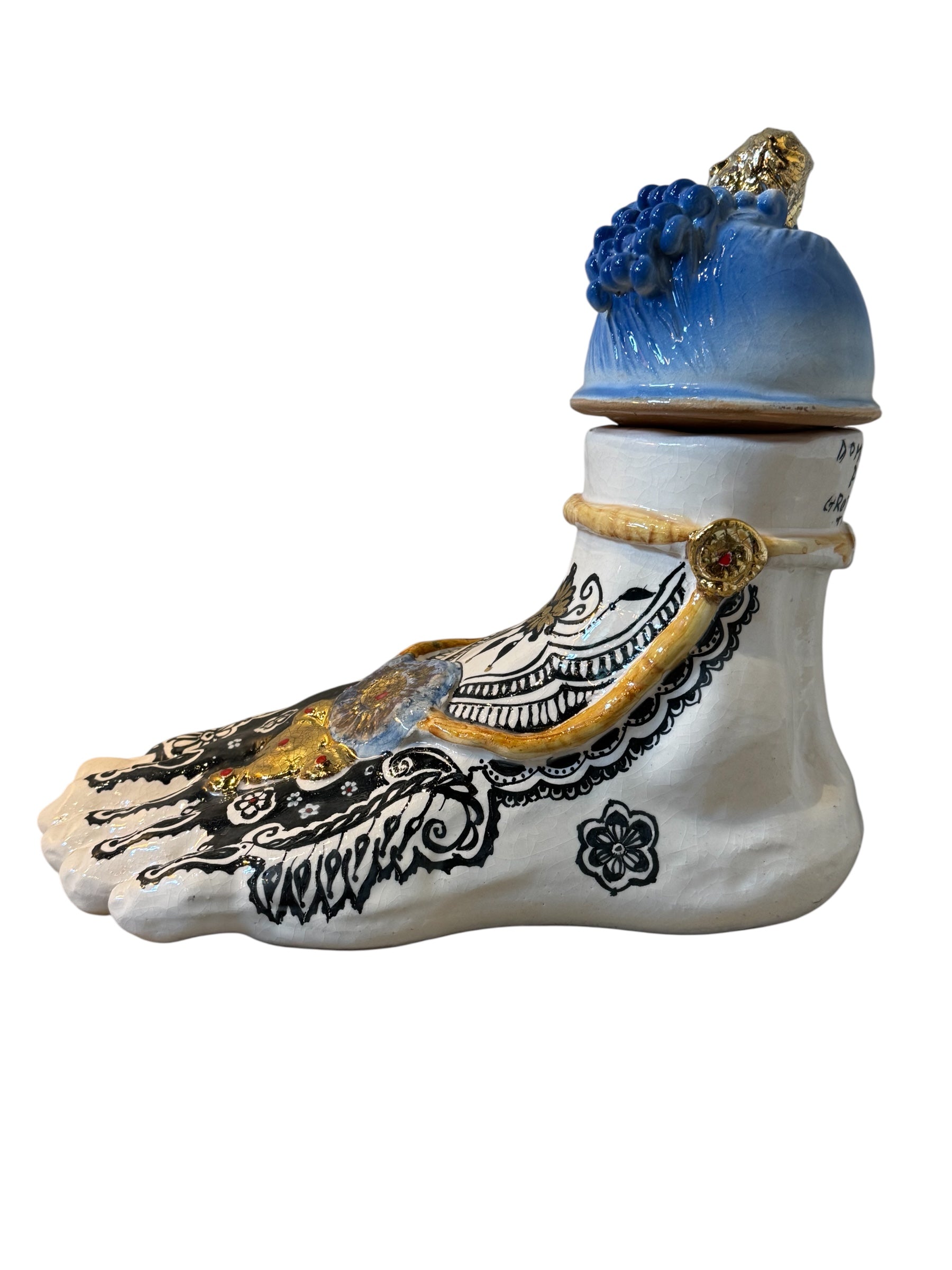 Handcrafted ceramic sculpture in the shape of a foot, decorated with intricate black and white patterns, 24KT gold accents, and topped with a blue lid featuring a gilt fish and head emerging from stylized waves.
