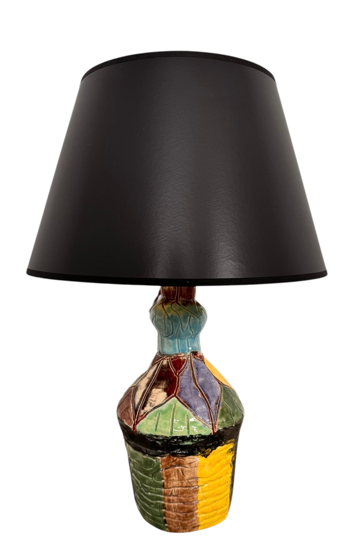 A hand-crafted ceramic table lamp featuring a vibrant, multi-colored base and a sleek black shade. The base is an upcycled artisan vase with a textured, hand-thrown appearance, decorated with geometric panels in shades of mustard yellow, forest green, cobalt blue, and burgundy, separated by thick black outlines. The lamp is topped with a minimalist, black tapered drum shade that provides a sharp contrast to the colorful, eclectic pottery below.