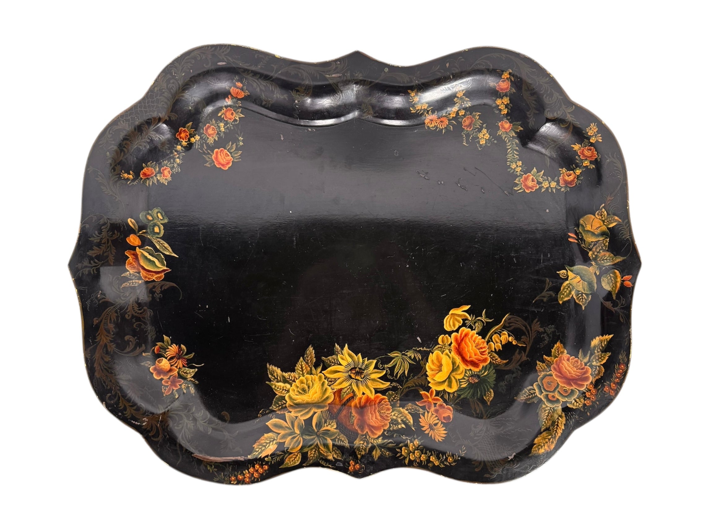 A vintage rectangular toleware serving tray with a scalloped, wavy edge and a black lacquered finish. The tray features hand-painted floral motifs in a traditional folk-art style. At the bottom center is a large, vibrant cluster of yellow and orange roses and daisies with green leaves. Smaller floral sprays and delicate gold filigree scrollwork decorate the corners and the upper rim. The center of the tray is open black space, showing light surface wear and minor scuffing consistent with age.