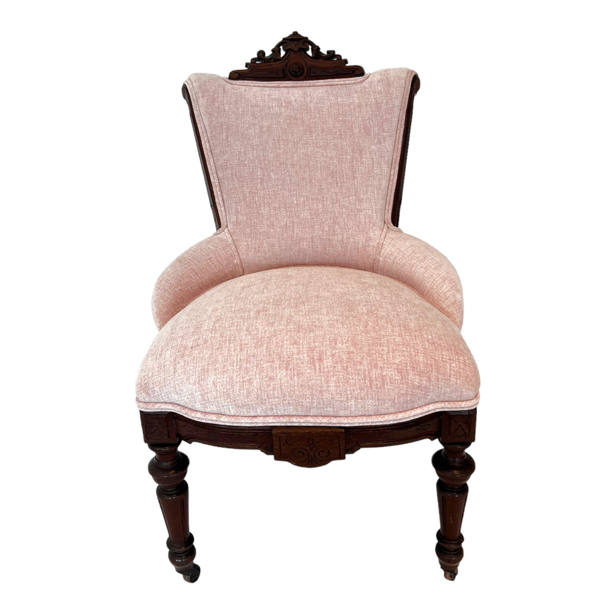 A hand carved Victorian parlor chair with a blush pink chenille upholstery and wood casters on the front legs.