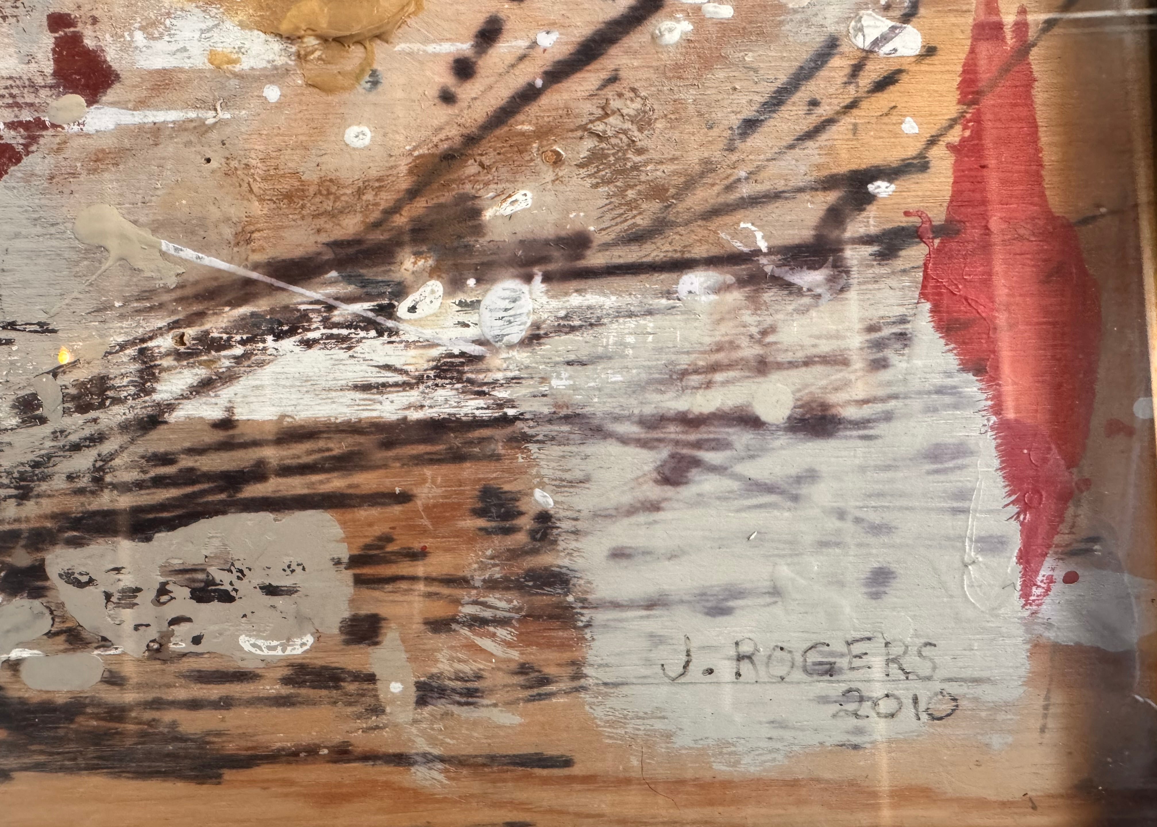 horizontal rectangular mixed media abstract artwork by J. Rogers, dated 2010, presented in a thin black frame. The composition is set against a light-toned wood grain background and features a dynamic, gestural arrangement of thick white impasto, splatters of red and black paint, and aggressive black scribbles. A vertical strip of textured material is collaged near the center, while various washes of tan and cream create a sense of weathered depth across the wooden surface.