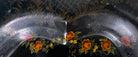 A vintage rectangular toleware serving tray with a scalloped, wavy edge and a black lacquered finish. The tray features hand-painted floral motifs in a traditional folk-art style. At the bottom center is a large, vibrant cluster of yellow and orange roses and daisies with green leaves. Smaller floral sprays and delicate gold filigree scrollwork decorate the corners and the upper rim. The center of the tray is open black space, showing light surface wear and minor scuffing consistent with age.