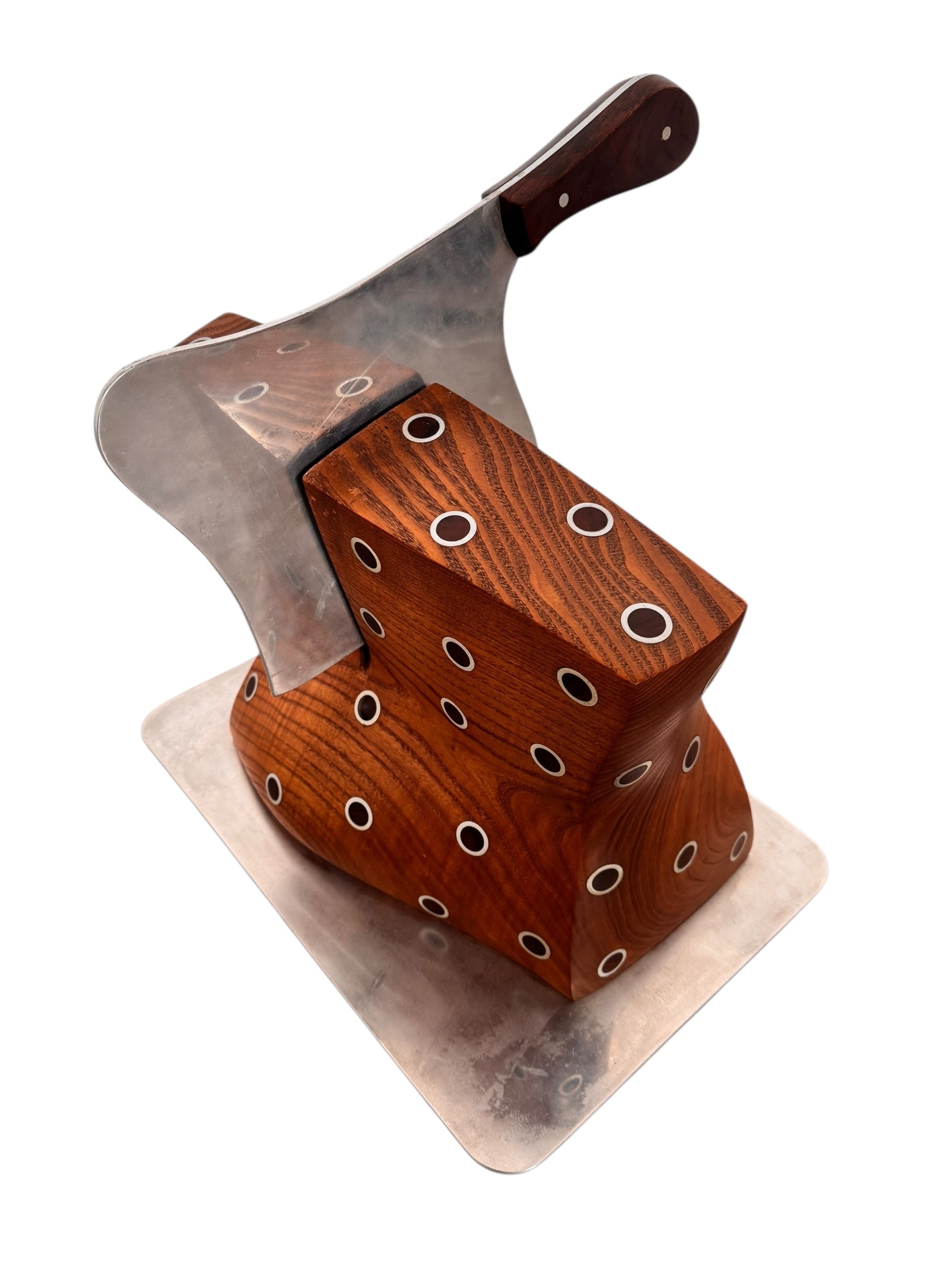 A wooden sculpture with a highly polished, reddish-brown grain, featuring two main asymmetrical, curved sections that converge at the top. Circular metal inlays with black centers are embedded across the surface of both wooden sections. A flat, polished metal blade-like element is inserted vertically between the two wooden sections, extending above the top surface. The sculpture rests on a metallic base.