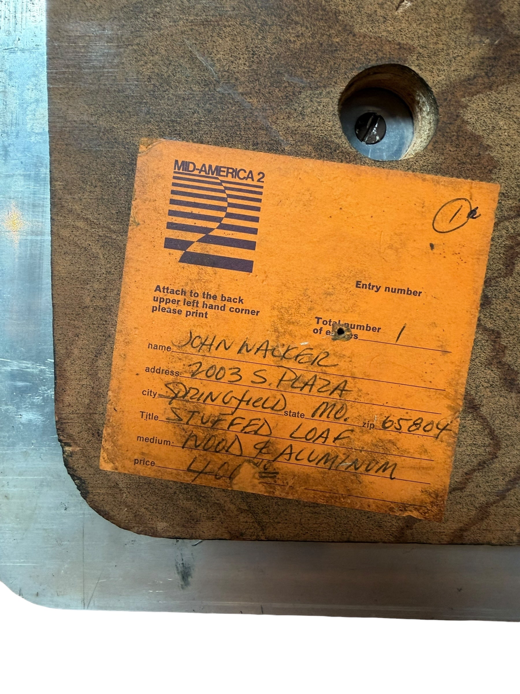 A wooden sculpture with a highly polished, reddish-brown grain, featuring two main asymmetrical, curved sections that converge at the top. Circular metal inlays with black centers are embedded across the surface of both wooden sections. A flat, polished metal blade-like element is inserted vertically between the two wooden sections, extending above the top surface. The sculpture rests on a metallic base.