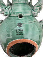 A ceramic sculpture of a stylized beetle by Enza Fasano, featuring a deep emerald green glaze with dark, metallic bronze-colored accents. The beetle is depicted with a highly textured, ribbed shell (elytra) and prominent, curved horns. Its legs are stylized with suckers, reminiscent of octopus tentacles. The piece has a glossy finish and rests against a plain white background.