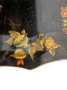 A vintage rectangular toleware serving tray with a scalloped, wavy edge and a black lacquered finish. The tray features hand-painted floral motifs in a traditional folk-art style. At the bottom center is a large, vibrant cluster of yellow and orange roses and daisies with green leaves. Smaller floral sprays and delicate gold filigree scrollwork decorate the corners and the upper rim. The center of the tray is open black space, showing light surface wear and minor scuffing consistent with age.