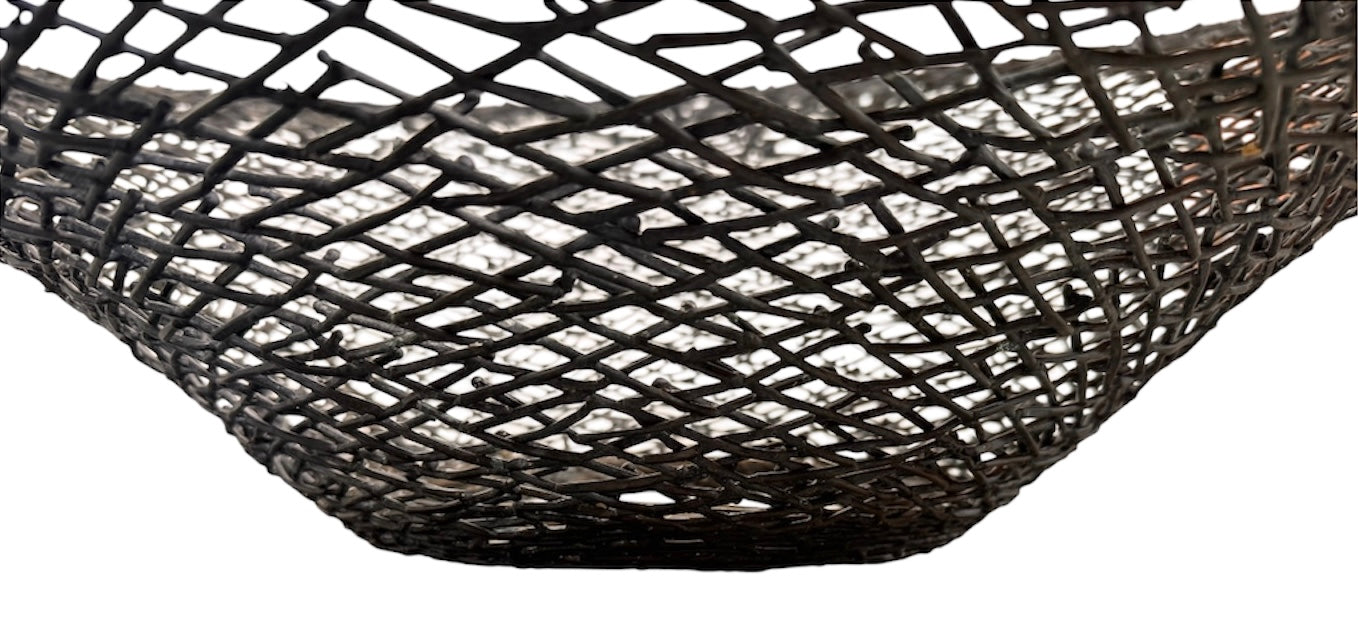 A round metal wall sculpture by Michael Aram, viewed from the front against a stark white background. The piece is composed of a dense, intricate lattice of fine, dark metal wire or twigs woven together to create a textured, web-like pattern. The structure curves inward like a funnel or a vortex, sloping down toward a clean, circular opening in the exact center, creating a strong sense of depth and dimensionality.