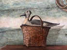 A realistic painting of a rustic interior scene by Travis Whitfield. A duck sits inside a woven basket on a dark wooden table with a single drawer. A French horn hangs on the light blue and white log wall behind the table. A multi-pane window on the left side of the painting shows an outdoor view. 
