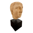 An ancient marble sculpted head of a young man, presented on a modern black presentation stand. The sculpture is rendered in a three-quarter view, angled slightly to the right, showing a classical profile characterized by a strong nose, softly contoured cheekbones, and slightly parted lips that convey a quiet, contemplative expression. The hair is carved in short, stylized strands that frame the forehead and ears, characteristic of ancient Roman or Greek portraiture styles.