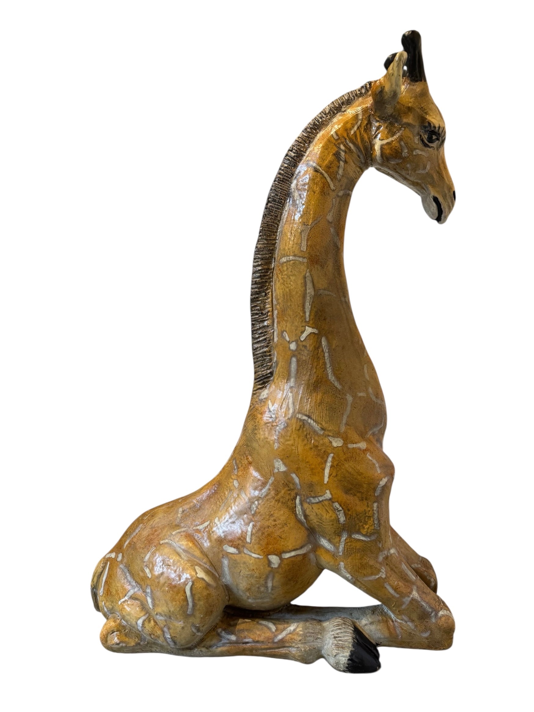 A hand-painted, cast resin sculpture of a seated giraffe. The giraffe has a long neck, is predominantly gold with light markings, and is in a resting pose. The sculpture is against a plain white background