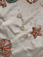 A round, cream-colored tablecloth featuring a dramatic, scalloped border shaped like six large flower petals. The border is intricately embroidered with a coastal motif including pink starfish, seashells, and teal-blue swirling waves. Gold beaded accents line the inner circular seam and the outer edges.
A long, narrow cream runner with matching scalloped, wavy edges. It is adorned with the same nautical embroidery of pink starfish and seashells scattered across the center and concentrated along the edge.