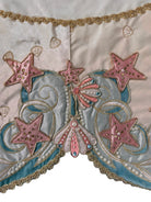 A round, cream-colored tablecloth featuring a dramatic, scalloped border shaped like six large flower petals. The border is intricately embroidered with a coastal motif including pink starfish, seashells, and teal-blue swirling waves. Gold beaded accents line the inner circular seam and the outer edges.
A long, narrow cream runner with matching scalloped, wavy edges. It is adorned with the same nautical embroidery of pink starfish and seashells scattered across the center and concentrated along the edge.