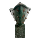curved ceramic piece resembling a green pod, supported by a block plinth. The seafoam-green ceramic structure extends wide on either side, curving upward to form a wavy, boat-like shape. The outer surface is dotted with rows of small, raised, brown-orange, acorn-like protrusions. The top rim is layered with parallel, grooved ridges. The base is a vertical rectangular block with a heavily textured, bark-like surface in a dark green and black finish.