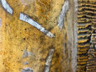 A hand-painted, cast resin sculpture of a seated giraffe. The giraffe has a long neck, is predominantly gold with light markings, and is in a resting pose. The sculpture is against a plain white background