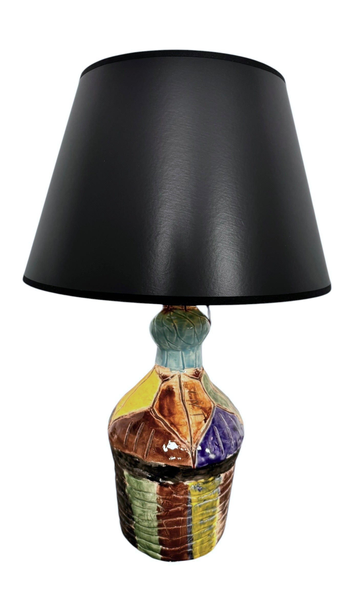 A hand-crafted ceramic table lamp featuring a vibrant, multi-colored base and a sleek black shade. The base is an upcycled artisan vase with a textured, hand-thrown appearance, decorated with geometric panels in shades of mustard yellow, forest green, cobalt blue, and burgundy, separated by thick black outlines. The lamp is topped with a minimalist, black tapered drum shade that provides a sharp contrast to the colorful, eclectic pottery below.
