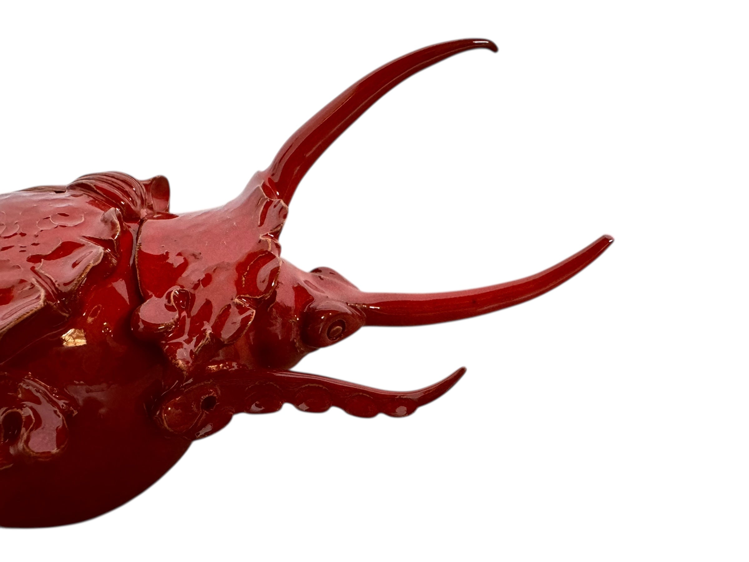 A handcrafted ceramic sculpture of a rhinoceros beetle by Enza Fasano, featuring a vibrant, glossy deep-red glaze. The piece is stylized with smooth, rounded contours and prominent, elegant horns extending forward. Its surface reflects light, highlighting the rich texture and artisanal detail of the clay.