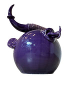 Glossy purple ceramic sculpture featuring a round body and stylized bull horns adorned with rings, and a nose ring. The sculpture has no eyes, but suggests a smiling expression.