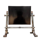 The antique Chevalier Mirror features a rectangular swing frame supported by two turned posts on decorative trestle feet. The mirror stands on four scrolled feet and features its original tarnished, silvered finish. The front of the mirror reflects a bright blue sky with wispy white clouds. The original dark brown backing is still attached to the frame.