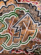 A close-up, angled view of an abstract oil painting titled 'Topography' by Robert Wymer. The painting's edge reveals thick, textured swirls of brown, gold, red, green, and white paint. It is framed in a natural light-brown teak wood frame, with a visible corner joint where two pieces of wood meet