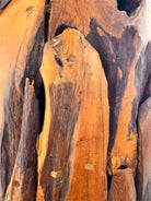 Two table lamps made from repurposed teak wood vases. The bases are tall and tapered, featuring a rugged, mosaic-like texture composed of various pieces of dark, polished teak wood with visible natural grain and crevices. Each lamp is topped with a large, warm-toned conical shade that is currently wrapped in protective clear plastic, emitting a soft, golden glow. The lamps are positioned on a clean white surface against a neutral background.
