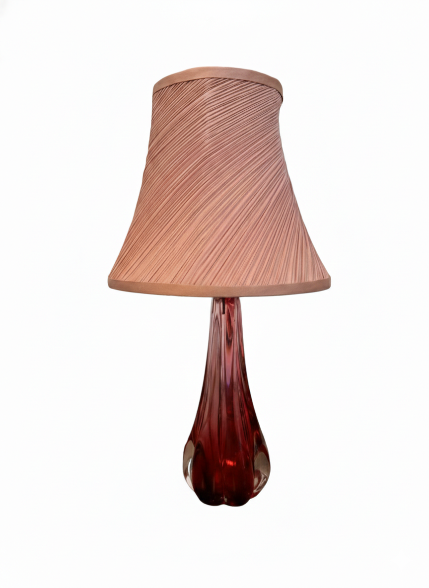 A table lamp featuring a teardrop-shaped, reddish-purple glass base and a conical, pleated, light-pink fabric shade.