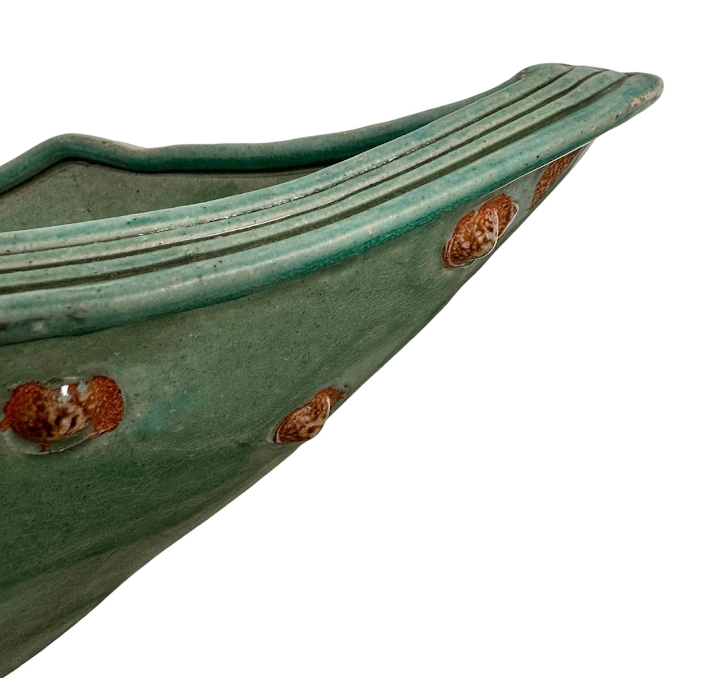 curved ceramic piece resembling a green pod, supported by a block plinth. The seafoam-green ceramic structure extends wide on either side, curving upward to form a wavy, boat-like shape. The outer surface is dotted with rows of small, raised, brown-orange, acorn-like protrusions. The top rim is layered with parallel, grooved ridges. The base is a vertical rectangular block with a heavily textured, bark-like surface in a dark green and black finish.