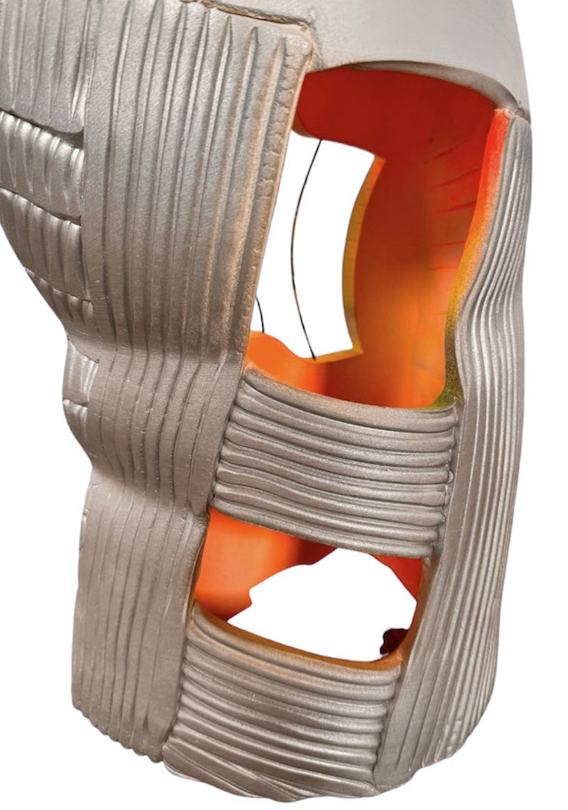 Carved, rib-like vertical lines cover the lower two-thirds of the form, with some sections resembling stacked bricks or tire treads. A subtle metallic silver finish catches the light on the right half, contrasting with the chalky finish on the left. The top third of the sculpture is a smooth, rounded cap. Rectangular cutouts on both sides pierce through the outer layer, revealing a hollow interior glowing with vibrant, airbrushed gradients of neon pink, orange, yellow, and green.