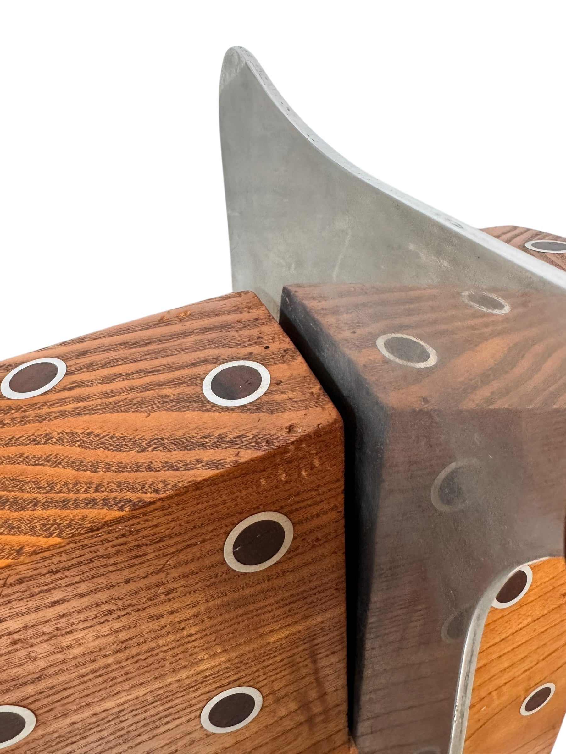 A wooden sculpture with a highly polished, reddish-brown grain, featuring two main asymmetrical, curved sections that converge at the top. Circular metal inlays with black centers are embedded across the surface of both wooden sections. A flat, polished metal blade-like element is inserted vertically between the two wooden sections, extending above the top surface. The sculpture rests on a metallic base.