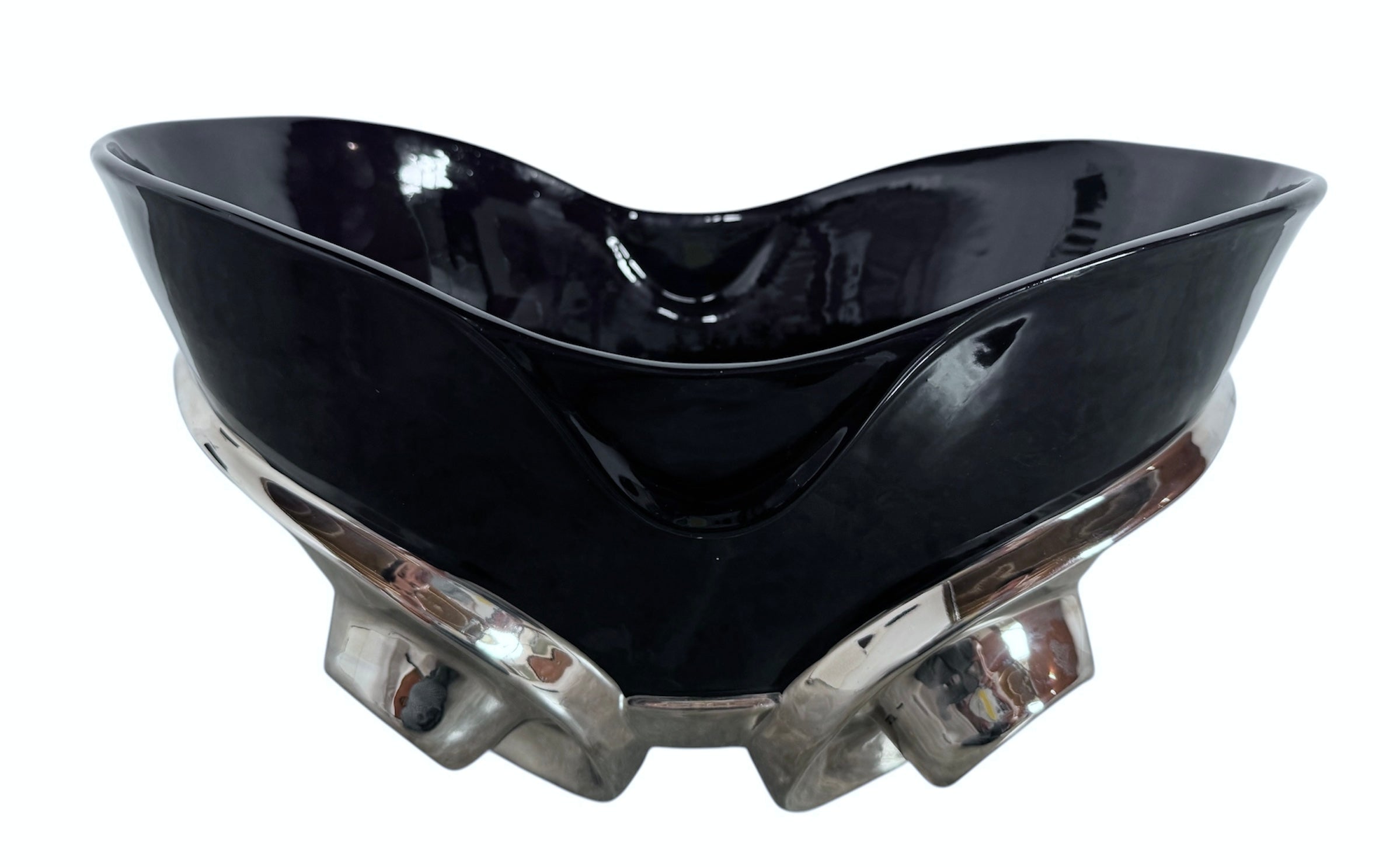 A large, decorative ceramic bowl by Dolbi Cashier in a dramatic Postmodern style. The top half is finished in a polished, piano-black glaze with an organic, wavy "butterfly" rim that dips in the center and rises at the sides. The bottom half is finished in a reflective, metallic silver chrome glaze, featuring bold, geometric-rounded feet and a structured base that appears to "hold" the black vessel. The overall shape is wide and sculptural, reminiscent of 1980s Art Deco Revival.