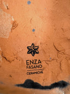 A dark, chocolate-brown ceramic sculpture by Enza Fasano depicting the stylized head of a hunting dog, such as a pointer or hound. The piece features a smooth, elongated snout and large, floppy ears with a textured, feathery finish that contrasts with the sleek crown of the head. The minimalist design emphasizes the dog's elegant profile and long neck.