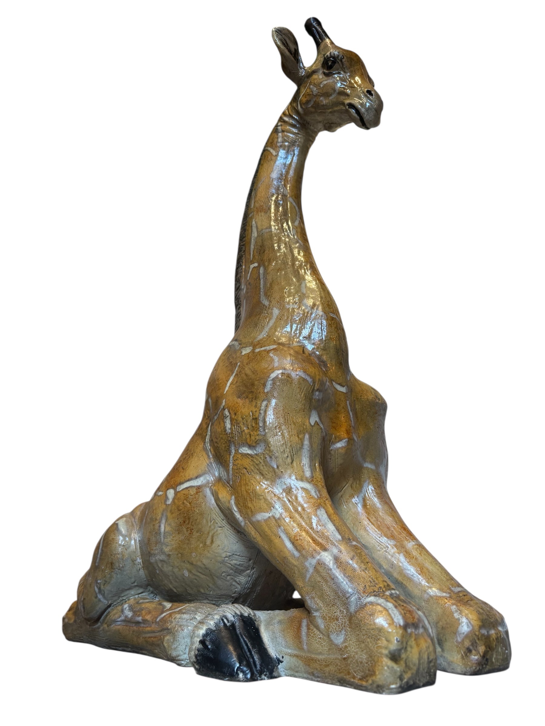 A hand-painted, cast resin sculpture of a seated giraffe. The giraffe has a long neck, is predominantly gold with light markings, and is in a resting pose. The sculpture is against a plain white background