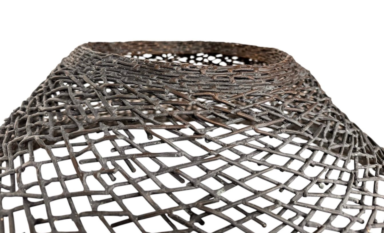 A round metal wall sculpture by Michael Aram, viewed from the front against a stark white background. The piece is composed of a dense, intricate lattice of fine, dark metal wire or twigs woven together to create a textured, web-like pattern. The structure curves inward like a funnel or a vortex, sloping down toward a clean, circular opening in the exact center, creating a strong sense of depth and dimensionality.