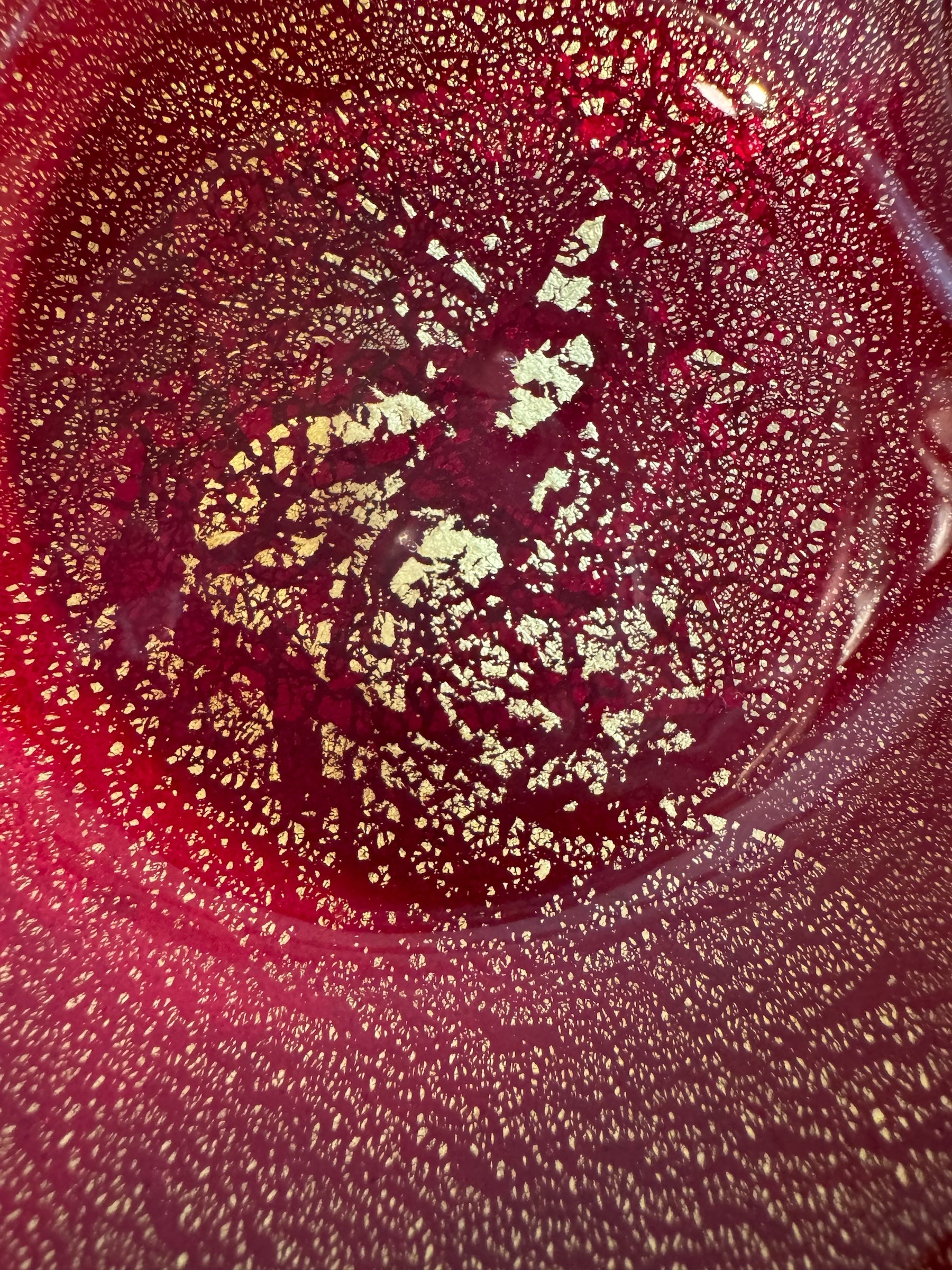 A handcrafted Murano glass bowl with a ruffled edge, deep red color, and gold specks created using the "oro zecchino" technique. The surface has a crackled gold pattern that reflects light, making it a striking decorative piece.