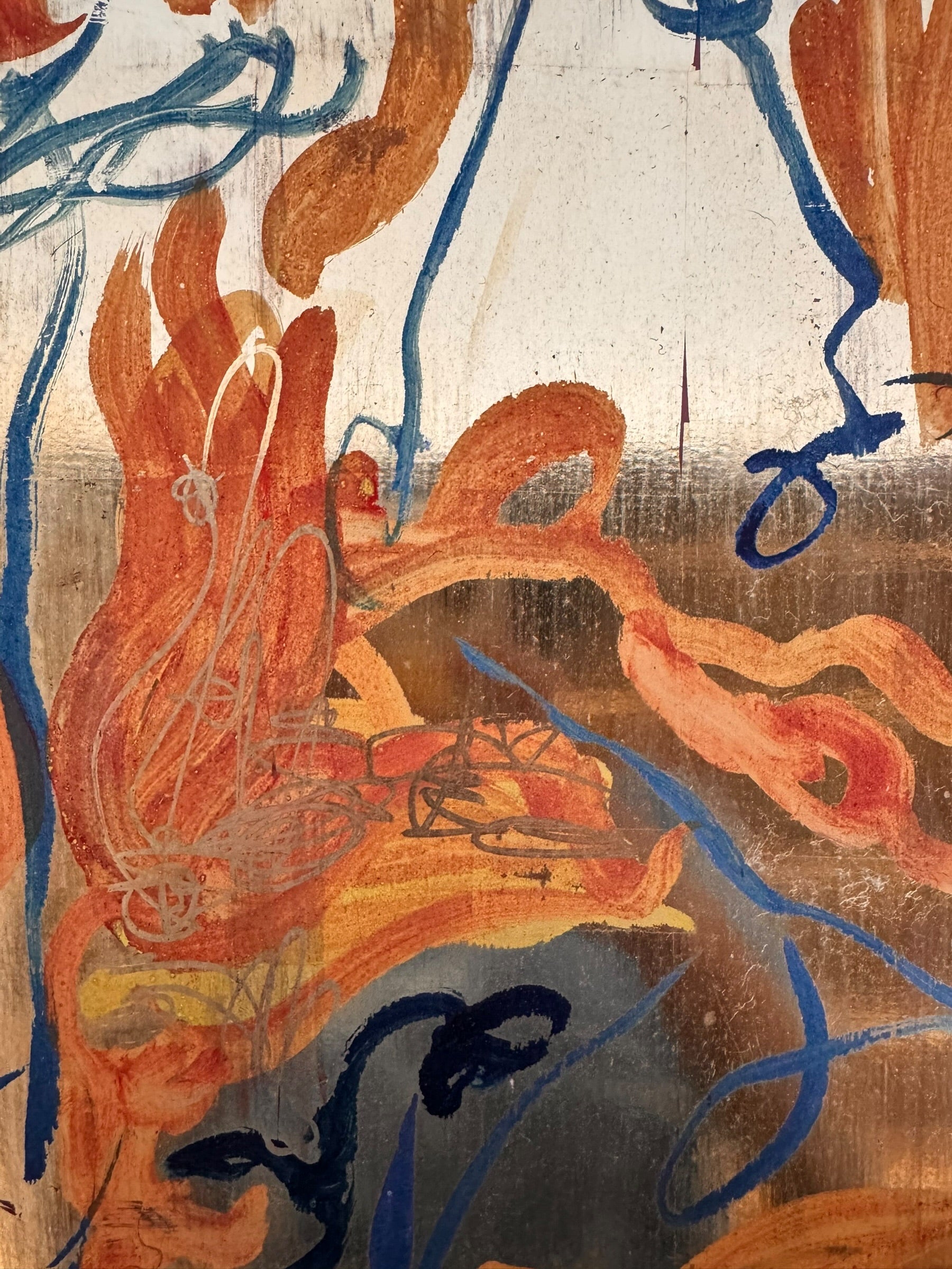 An abstract mixed-media painting on a vertical rectangular panel with a reflective, metallic gold and copper background. The composition features fluid, expressive brushstrokes in warm orange, ochre, and deep blue.
The orange strokes are thick and organic, resembling swirling flames or floral petals, while the blue marks consist of thin, lyrical lines and looped scribbles that dance across the surface. The metallic substrate is visible through the translucent paint, creating a play of light and texture
