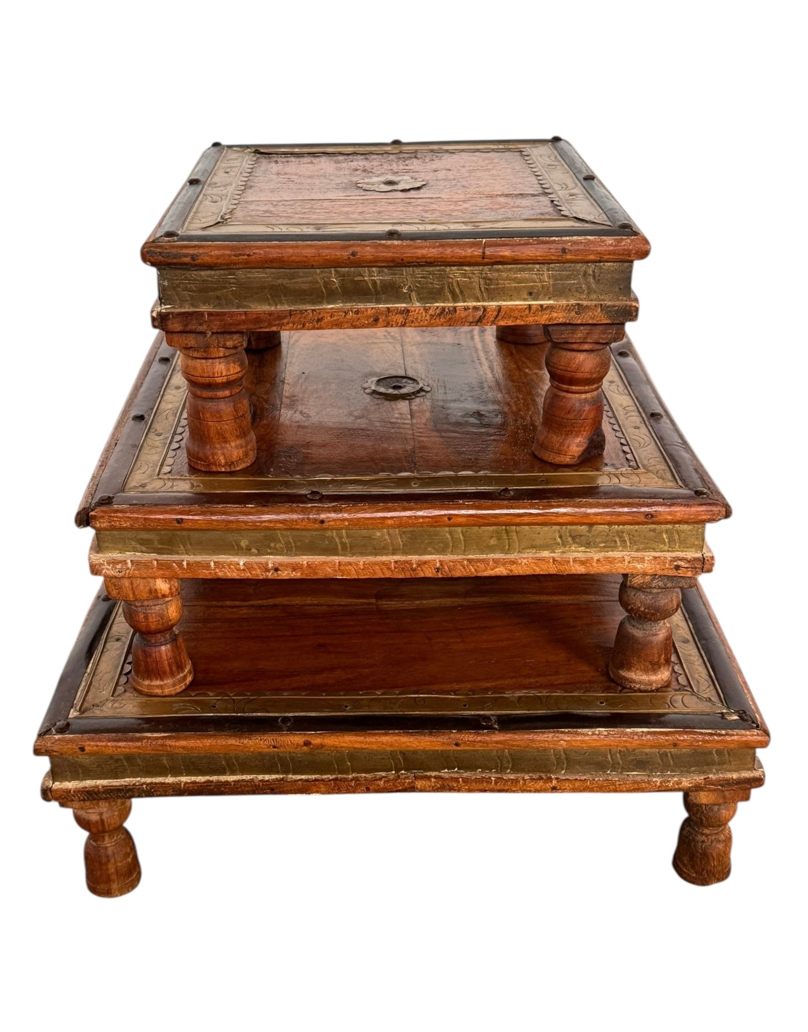 Stack of three vintage Indian teak bajot nesting tables featuring brass-clad edges, repoussé details, and short turned legs against a white background.