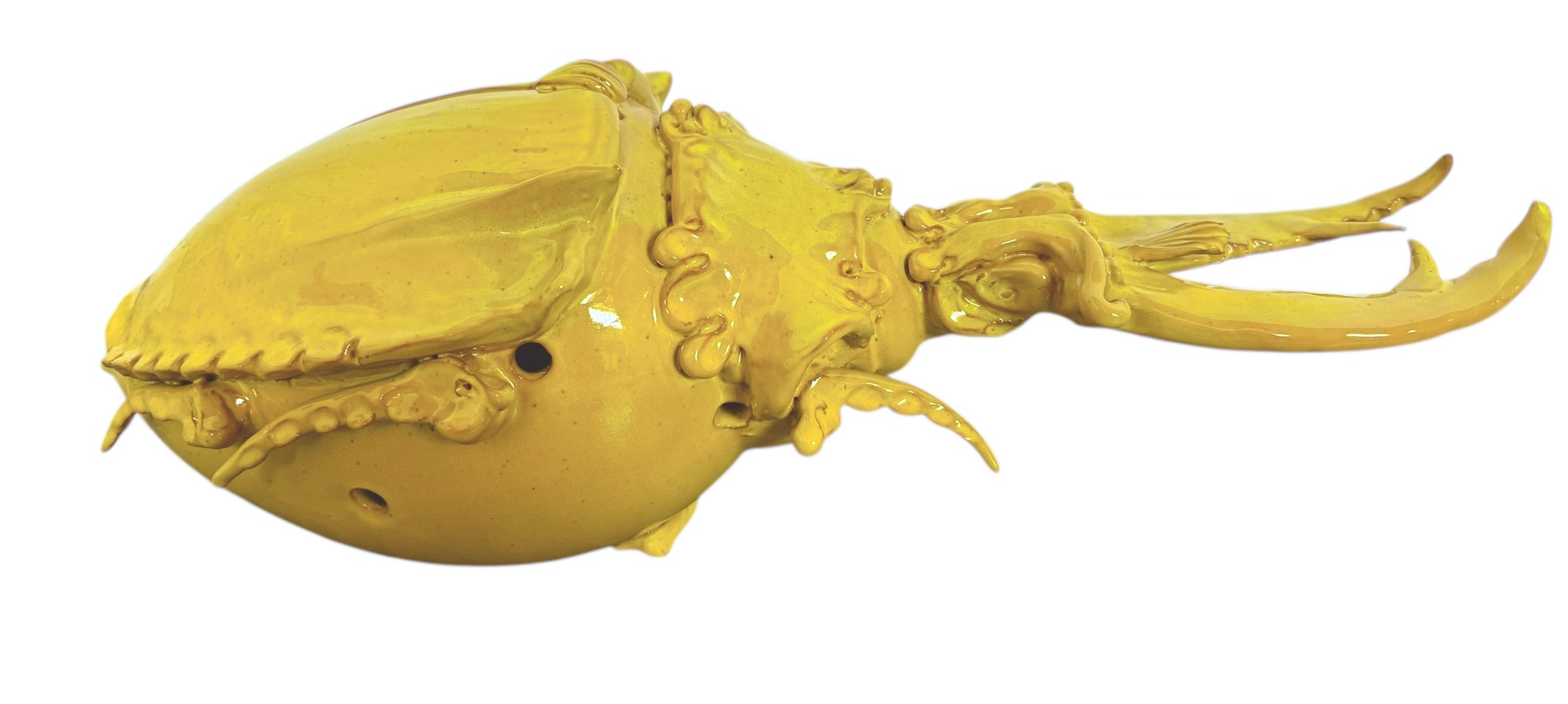 a large, high-gloss ceramic sculpture by Enza Fasano, shaped like a stylized beetle. The entire piece is finished in a warm, honey-yellow glaze. The beetle features prominent, curved mandibles at the front, a textured "collar" area, and a rounded, shell-like body with decorative ridges along the edges. The surface is smooth and reflective, catching the light across its organic contours.