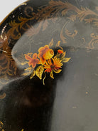 A vintage rectangular toleware serving tray with a scalloped, wavy edge and a black lacquered finish. The tray features hand-painted floral motifs in a traditional folk-art style. At the bottom center is a large, vibrant cluster of yellow and orange roses and daisies with green leaves. Smaller floral sprays and delicate gold filigree scrollwork decorate the corners and the upper rim. The center of the tray is open black space, showing light surface wear and minor scuffing consistent with age.
