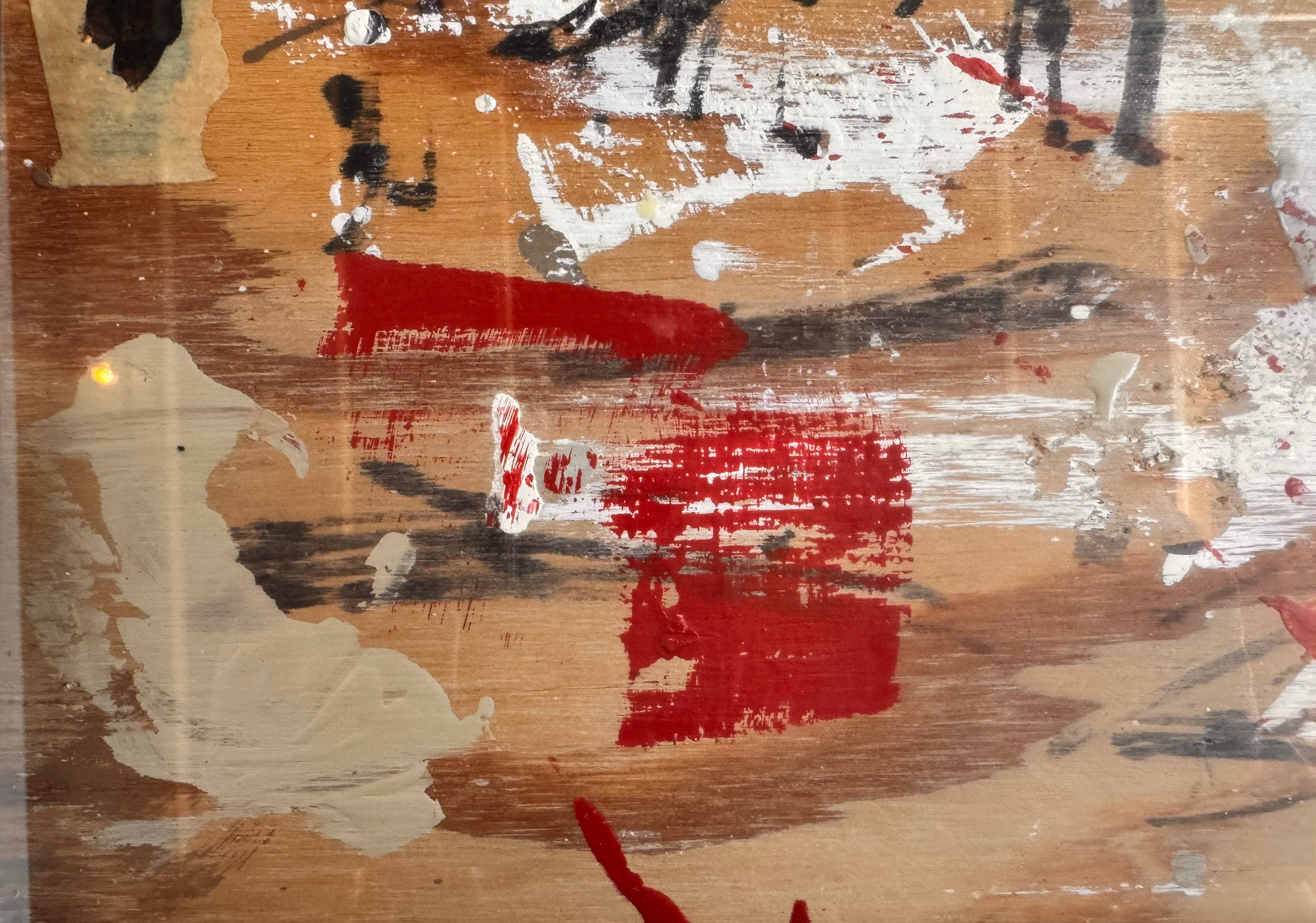 horizontal rectangular mixed media abstract artwork by J. Rogers, dated 2010, presented in a thin black frame. The composition is set against a light-toned wood grain background and features a dynamic, gestural arrangement of thick white impasto, splatters of red and black paint, and aggressive black scribbles. A vertical strip of textured material is collaged near the center, while various washes of tan and cream create a sense of weathered depth across the wooden surface.