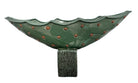 curved ceramic piece resembling a green pod, supported by a block plinth. The seafoam-green ceramic structure extends wide on either side, curving upward to form a wavy, boat-like shape. The outer surface is dotted with rows of small, raised, brown-orange, acorn-like protrusions. The top rim is layered with parallel, grooved ridges. The base is a vertical rectangular block with a heavily textured, bark-like surface in a dark green and black finish.