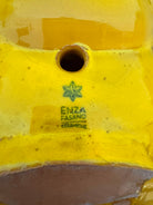 a large, high-gloss ceramic sculpture by Enza Fasano, shaped like a stylized beetle. The entire piece is finished in a warm, honey-yellow glaze. The beetle features prominent, curved mandibles at the front, a textured "collar" area, and a rounded, shell-like body with decorative ridges along the edges. The surface is smooth and reflective, catching the light across its organic contours.