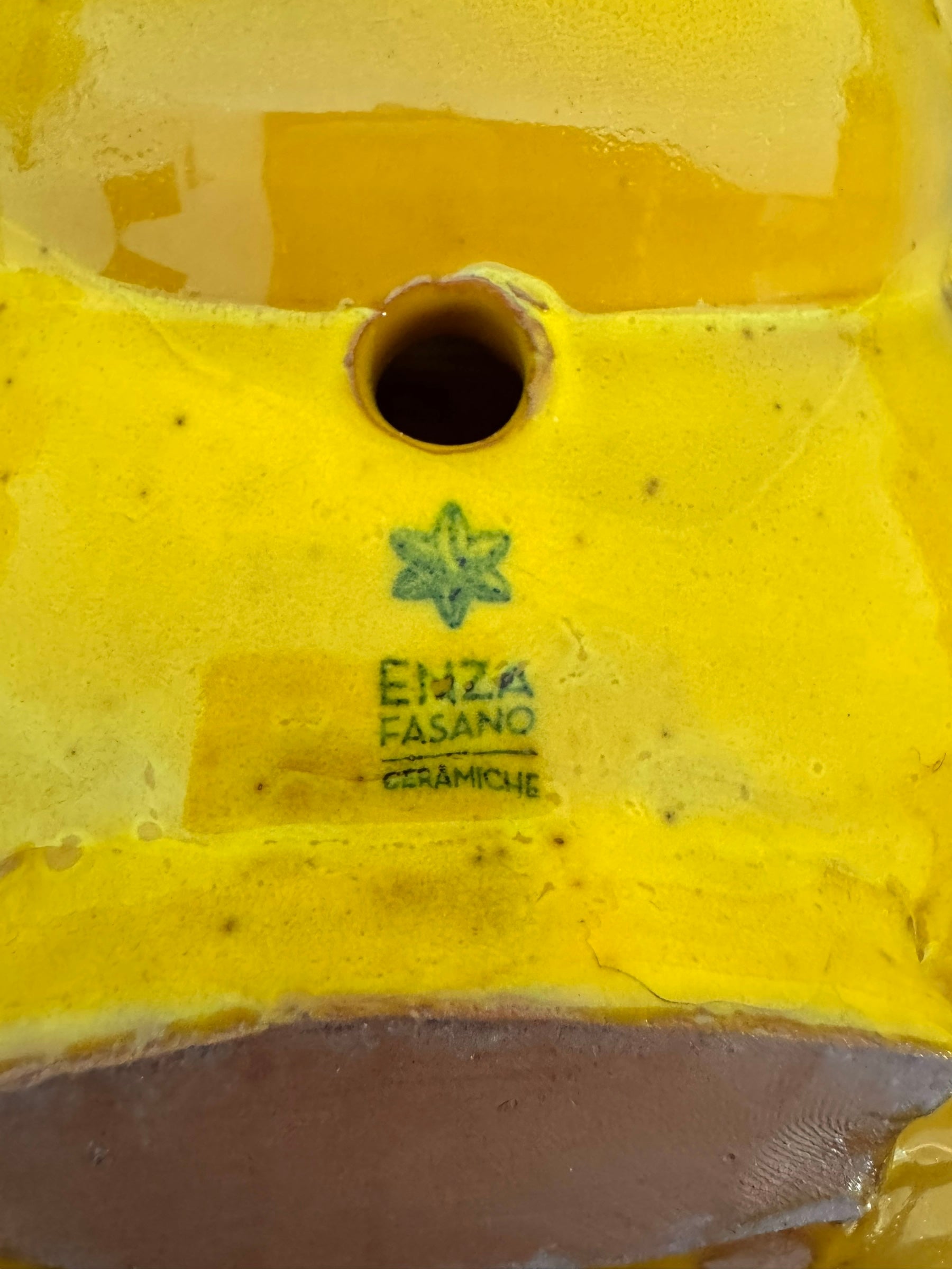 a large, high-gloss ceramic sculpture by Enza Fasano, shaped like a stylized beetle. The entire piece is finished in a warm, honey-yellow glaze. The beetle features prominent, curved mandibles at the front, a textured "collar" area, and a rounded, shell-like body with decorative ridges along the edges. The surface is smooth and reflective, catching the light across its organic contours.