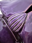 A glossy, deep purple ceramic sculpture of a beetle by Enza Fasano. The piece features a highly stylized and ornate design, blending insect-like anatomy with fantastical elements. It has a smooth, rounded shell and elongated, curved mandibles and appendages lined with sharp, thorn-like protrusions. The monochromatic glaze is reflective, highlighting the intricate sculptural details and textures of the avant-garde artwork.