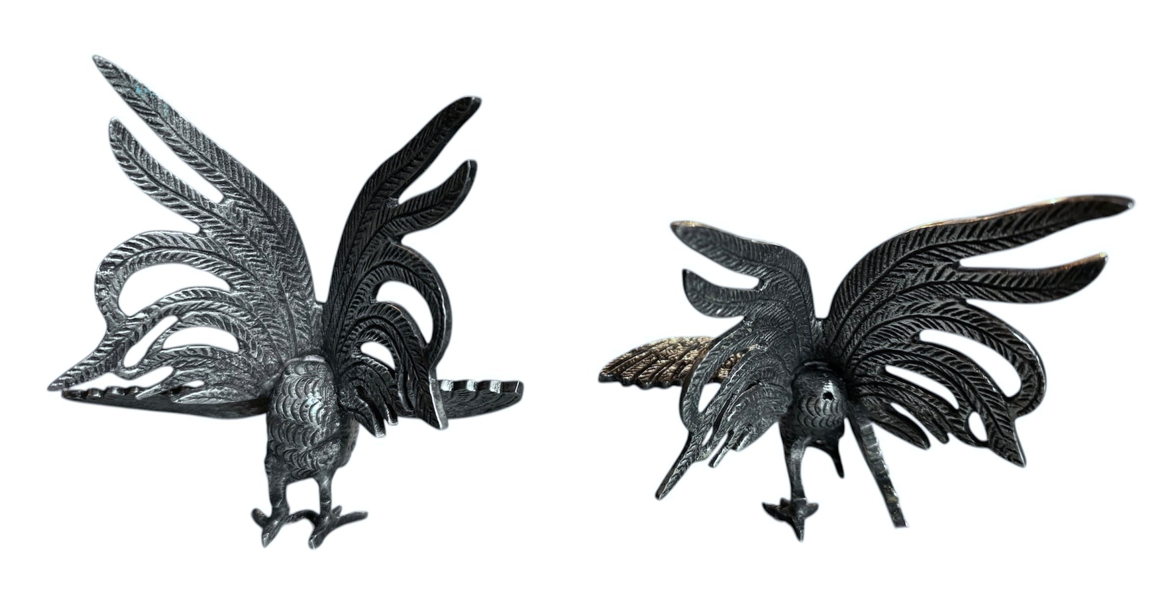 A pair of vintage, silver-plated brass fighting rooster figurines positioned in a confrontational stance against a white background. The rooster on the left is crouched low with its head down and tail feathers fanned high, while the rooster on the right stands more upright with its wings slightly flared. Both feature intricate feather detailing, a metallic patina showing hints of brass through the silver, and small, bright red glass eyes.