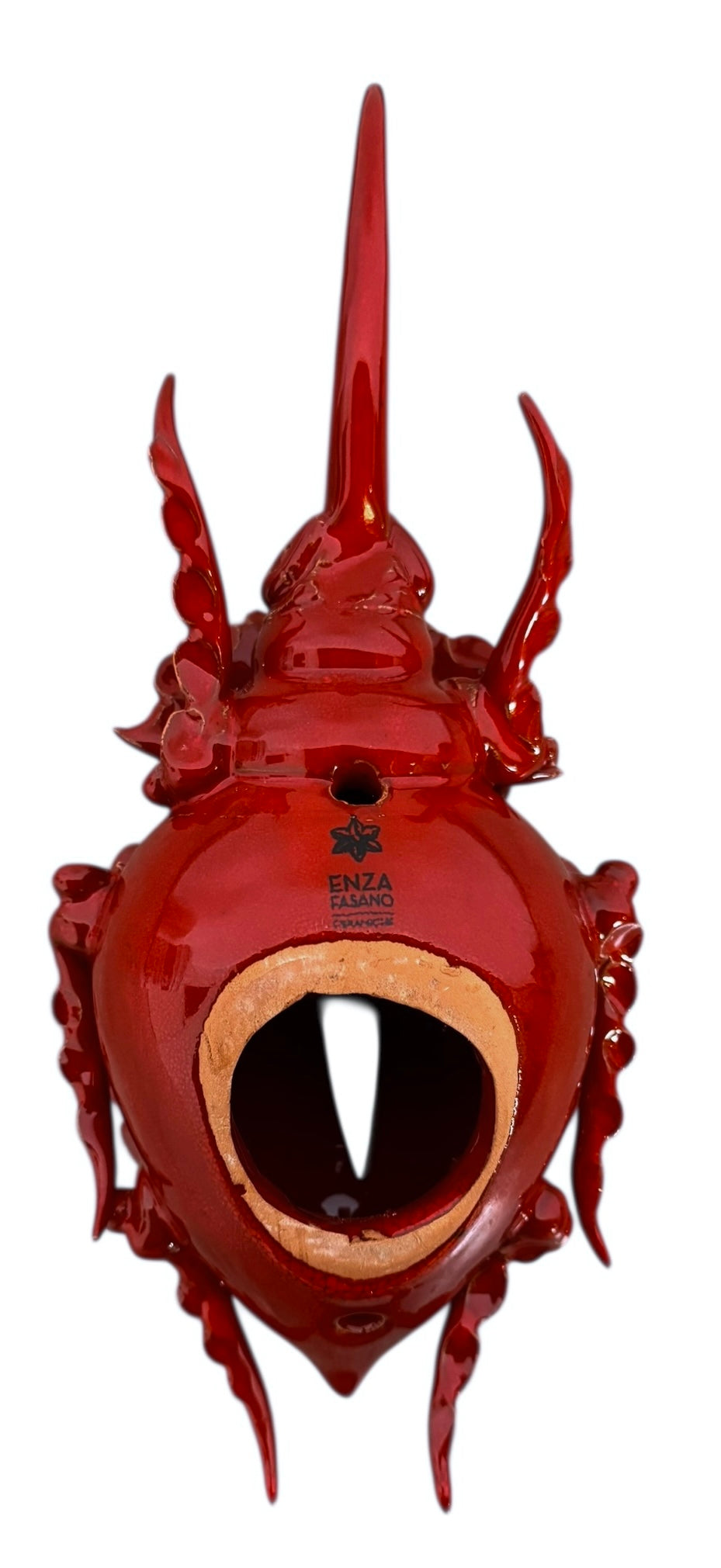 A handcrafted ceramic sculpture of a rhinoceros beetle by Enza Fasano, featuring a vibrant, glossy deep-red glaze. The piece is stylized with smooth, rounded contours and prominent, elegant horns extending forward. Its surface reflects light, highlighting the rich texture and artisanal detail of the clay.