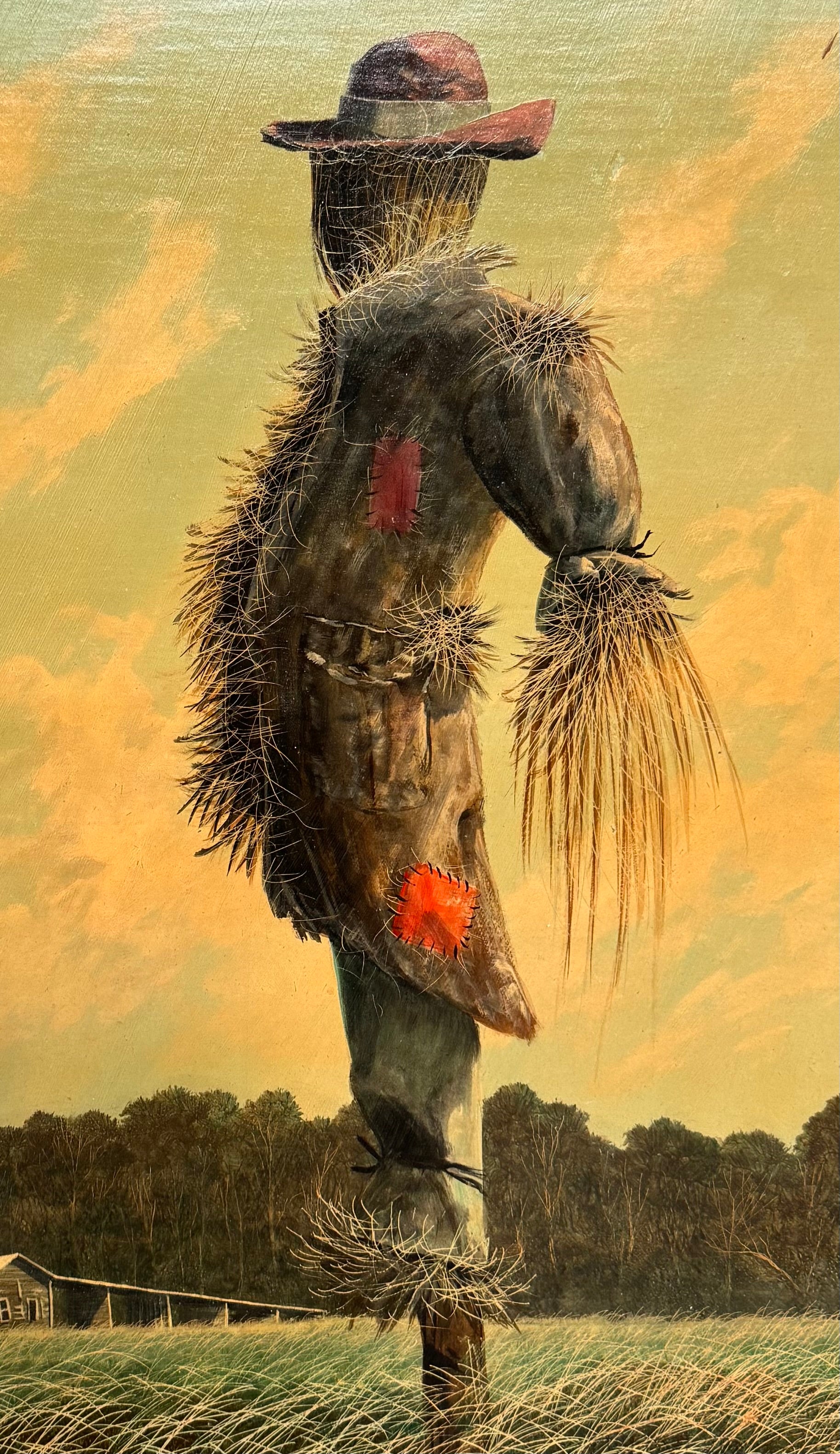 An oil painting on a wooden panel depicts a solitary scarecrow standing in a vast, grassy field under a pale yellow and blue sky. The scarecrow is shown from a three-quarters back view, wearing a brimmed hat and a tattered, patched brown coat with frayed straw bursting from its sleeves and edges. In the far left background, a long, white rustic barn sits before a dense line of green trees. The entire piece is enclosed in a thick, weathered, natural wood frame.