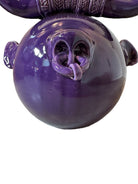 Glossy purple ceramic sculpture featuring a round body and stylized bull horns adorned with rings, and a nose ring. The sculpture has no eyes, but suggests a smiling expression.
