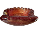 A handcrafted Murano glass bowl with a ruffled edge, deep red color, and gold specks created using the "oro zecchino" technique. The surface has a crackled gold pattern that reflects light, making it a striking decorative piece.