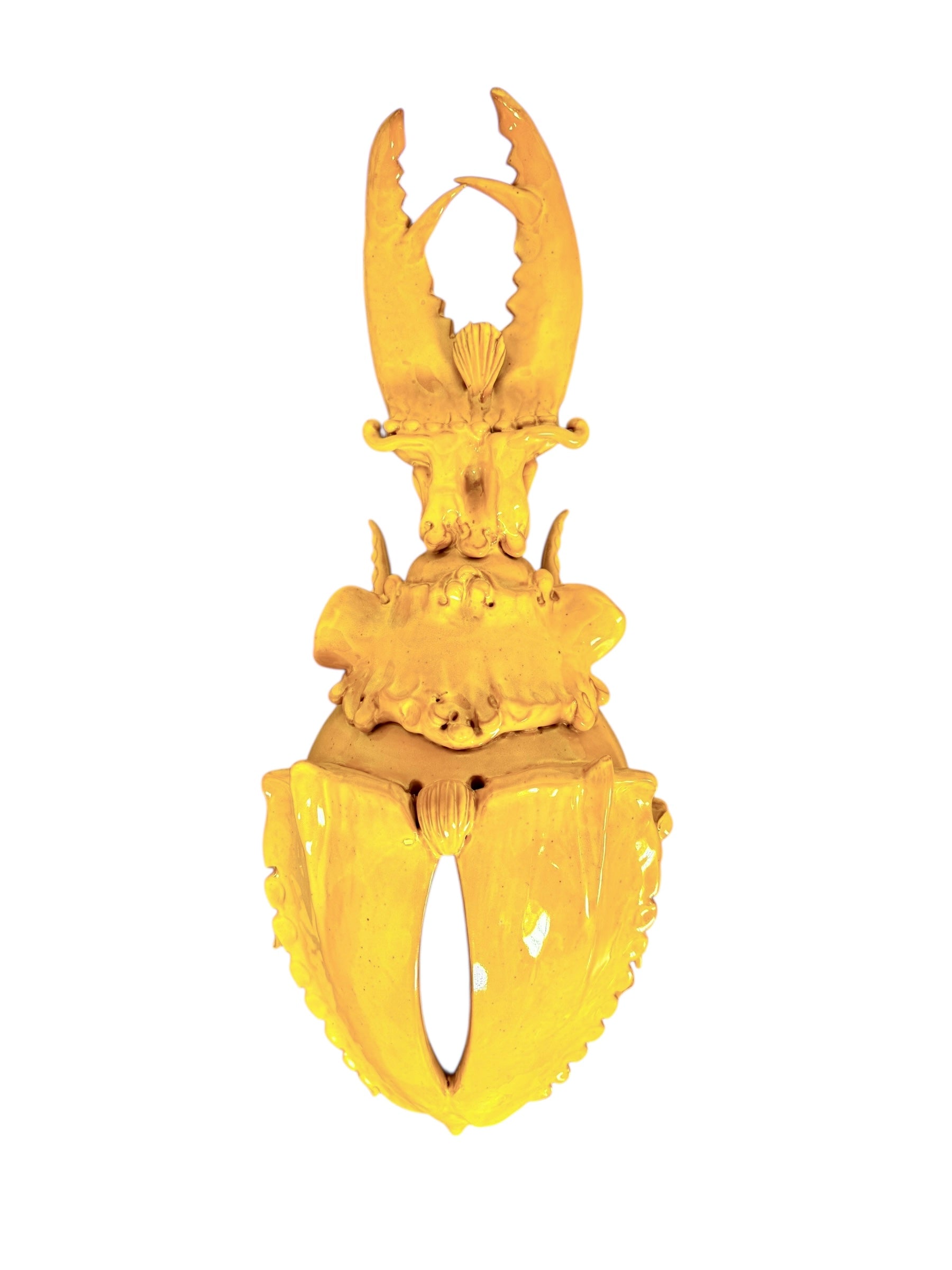 a large, high-gloss ceramic sculpture by Enza Fasano, shaped like a stylized beetle. The entire piece is finished in a warm, honey-yellow glaze. The beetle features prominent, curved mandibles at the front, a textured "collar" area, and a rounded, shell-like body with decorative ridges along the edges. The surface is smooth and reflective, catching the light across its organic contours.